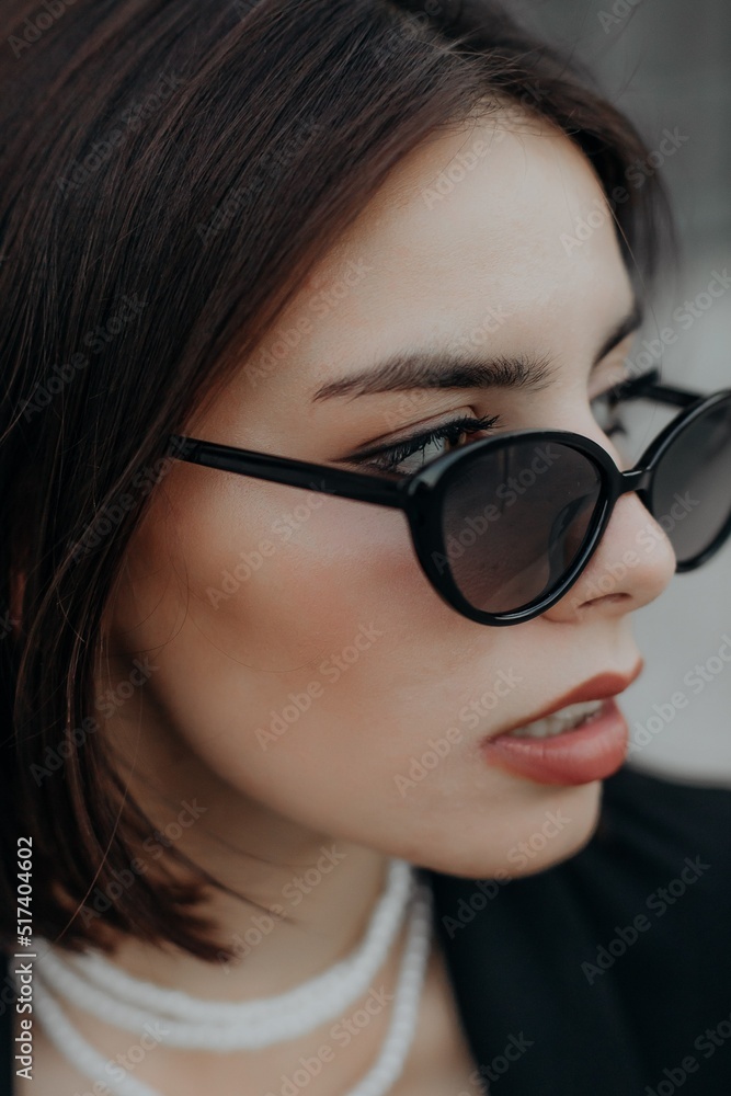 a brunette girl in a black jacket and dress and black glasses sits in the city with a handbag a brunette girl in a black jacket and dress and black glasses sits in the city with a handbag