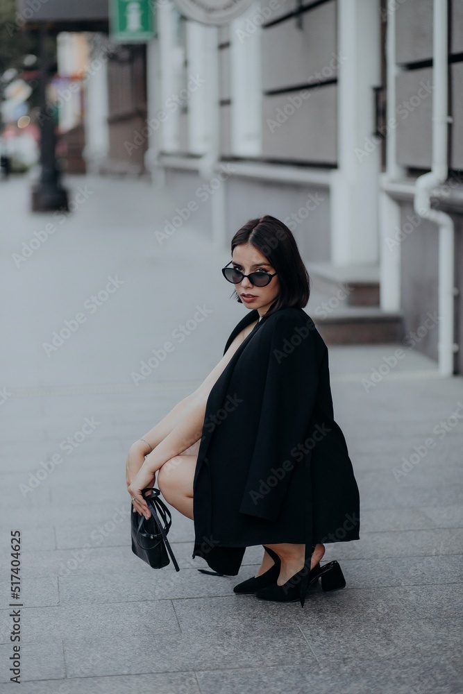 a brunette girl in a black jacket and dress and black glasses sits in the city with a handbag a brunette girl in a black jacket and dress and black glasses sits in the city with a handbag