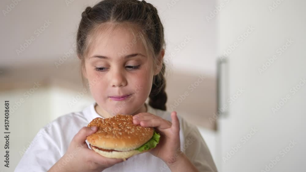 little girl eating a hamburger. unhealthy fast food proper nutrition concept. child greedily with pleasure bites a lifestyle big burger in the kitchen at home. kid eats fast food close-up little girl eating a hamburger. unhealthy fast food proper nutrition concept. child greedily with pleasure bites a lifestyle big burger in the kitchen at home. kid eats fast food close-up