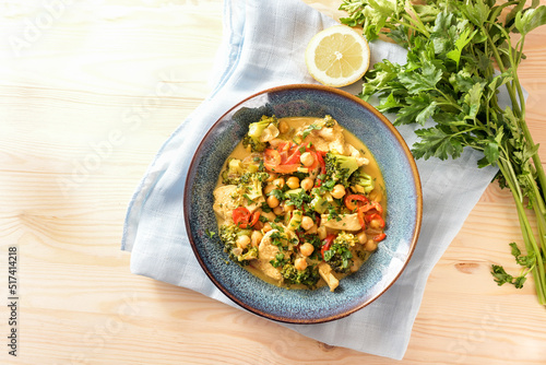 Indian style curry from chickpeas, colorful vegetables and chicken meat in a blue bowl, lemon, herbs and napkin on a light wooden table, copy space, high angle view