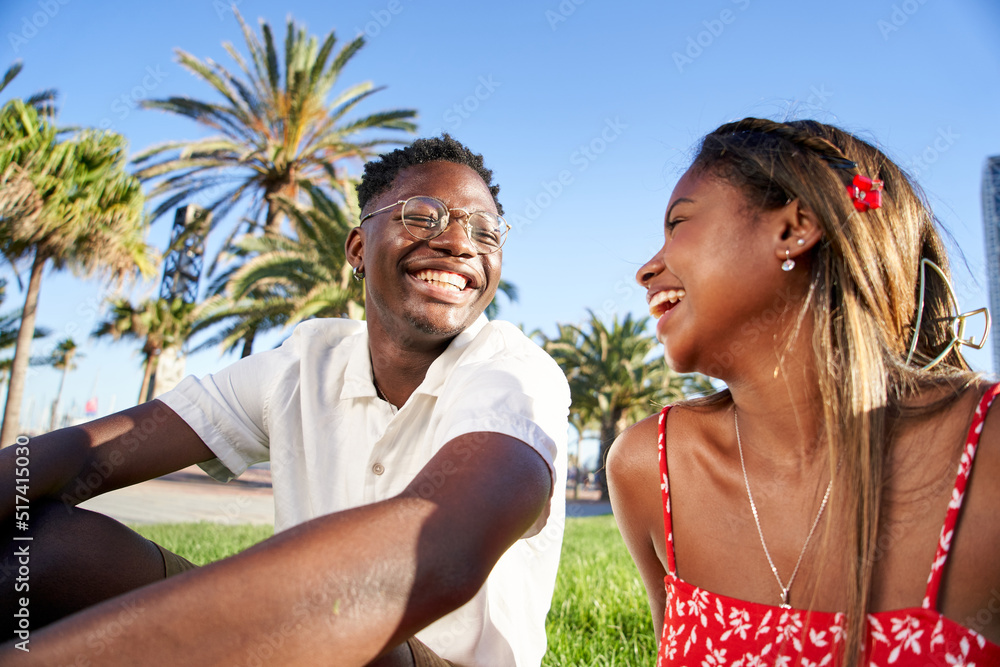 Obraz premium Cheerful young African couple laughing and having fun on their first date in the park in a romantic outdoor lovers scene, sitting on the grass talking