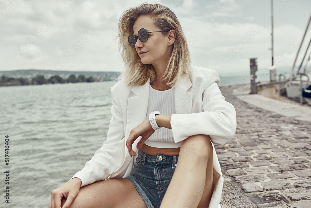 Fashion portrait of beautiful blondy woman sitting near the sea. White stylish blazer , sunglasses and watches. Modern clothes.