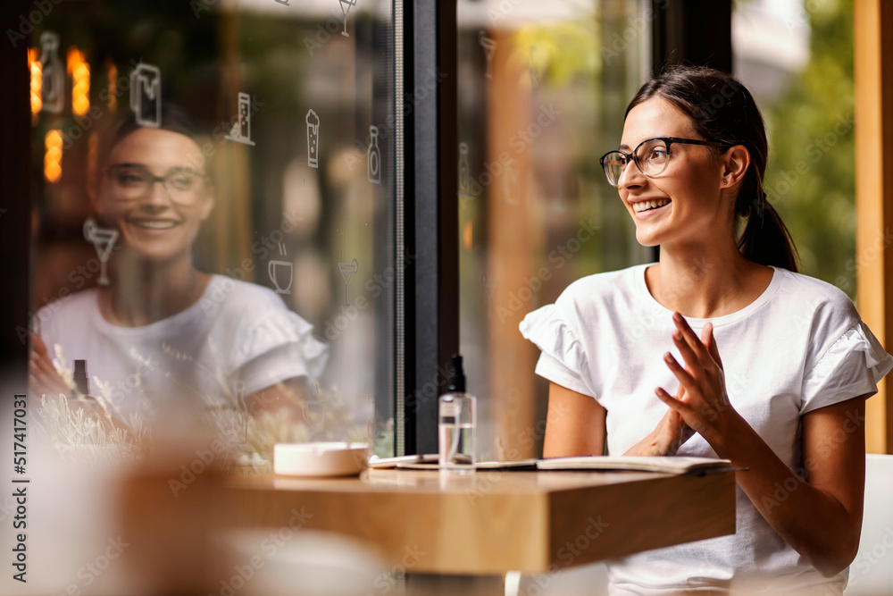 A happy young woman is sitting in a bar, disinfecting her hands and looking through the window. A happy young woman is sitting in a bar, disinfecting her hands and looking through the window.