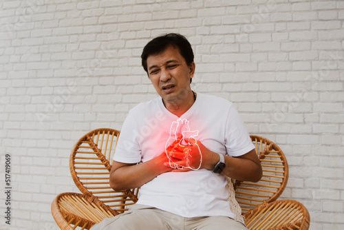 Middle aged, senior Asian man with sudden onset of heart disease showing pain heart muscle leaks tired and suffocating sits on a wooden chair in agony : Myocardial infarction (Heart Attack) photo