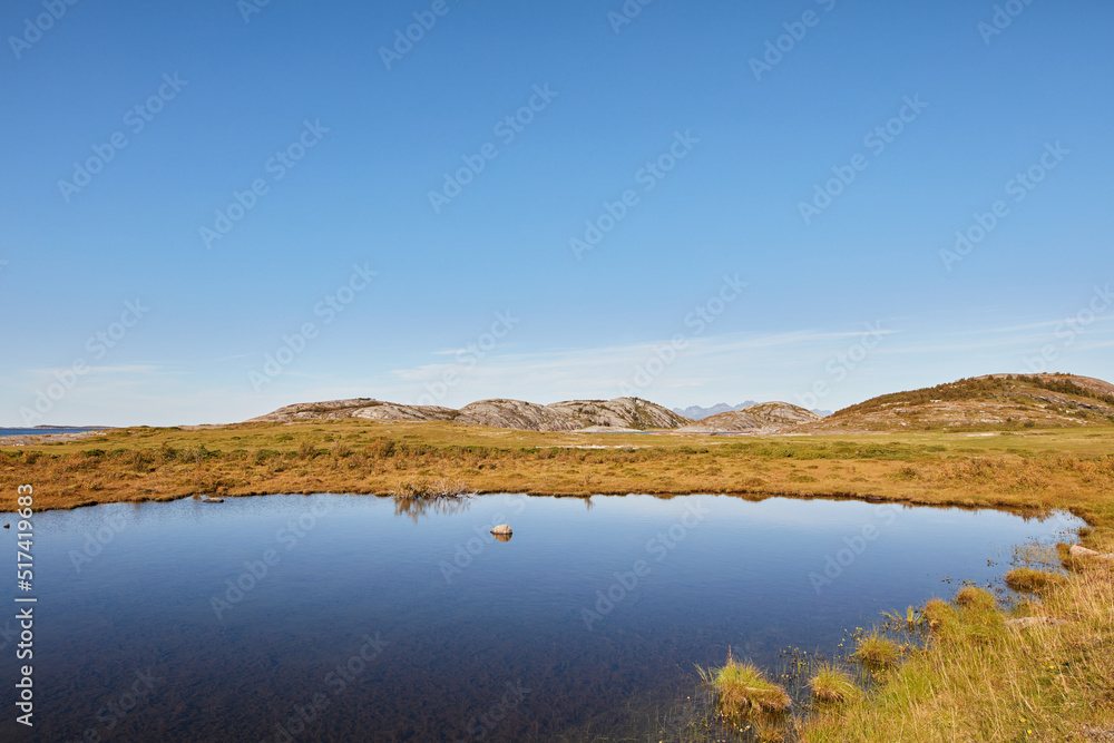 Landscape of a lake near grass fields against a blue horizon with mountain hills. Calm water in marsh land on a sunny day near Bodo in Norway. Peaceful and secluded fishing location in scenic nature