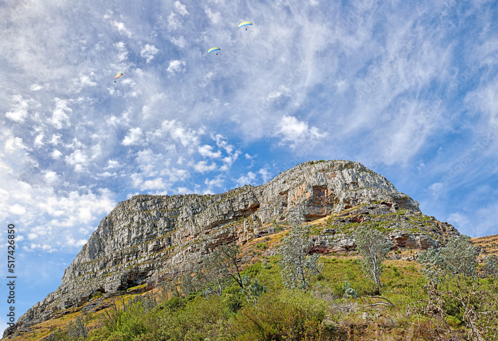 A photo of Lions Head and surroundings in Cape Town. Backdrop of rocky mountain with green vegetation and trees against blue sky filled with small wispy clouds. Barren landscape of natural wonder
