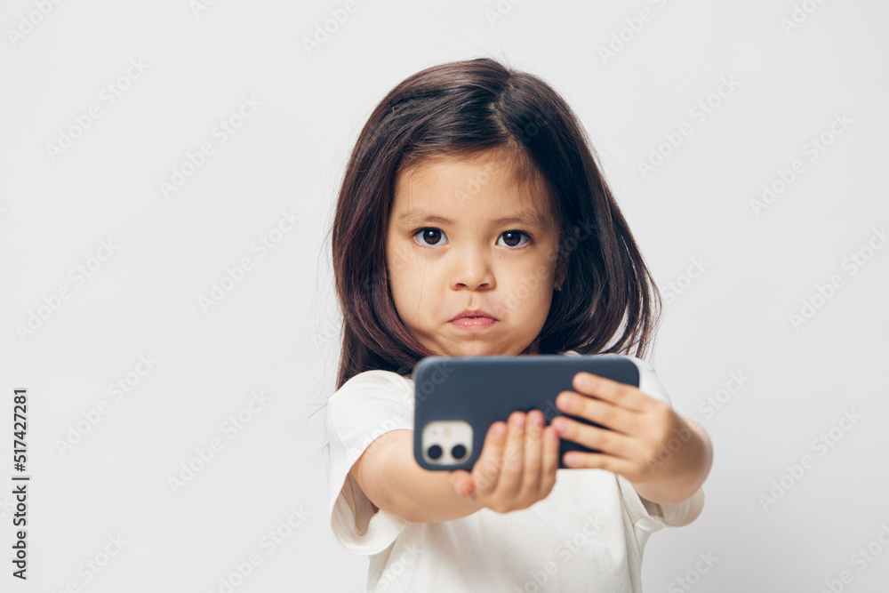 a cute little preschool girl stands on a white background in a white T-shirt and takes pictures of herself on her phone, smiling happily at the camera. The theme of children's happiness a cute little preschool girl stands on a white background in a white T-shirt and takes pictures of herself on her phone, smiling happily at the camera. The theme of children's happiness