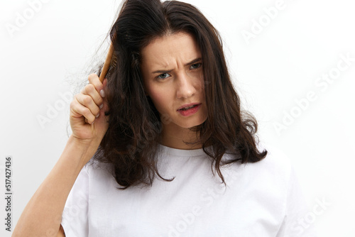 a sad, upset woman tries to comb her long, dark, tangled hair with a wooden massage comb, standing in a white T-shirt on a white background