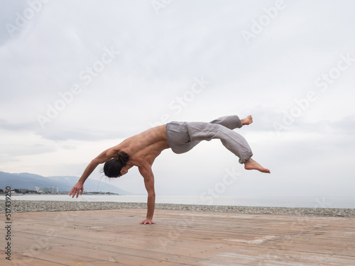 Shirtless caucasian man doing backflip on pebble beach. 