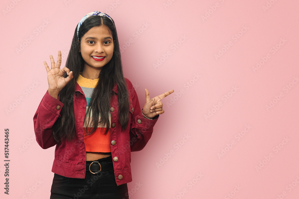 Young Indian Asian smiling cute girl posing isolated with one hand showing ok sign assuring you, confirm something is good and other directing towards copyspace showing recommendation being impressed Young Indian Asian smiling cute girl posing isolated with one hand showing ok sign assuring you, confirm something is good and other directing towards copyspace showing recommendation being impressed
