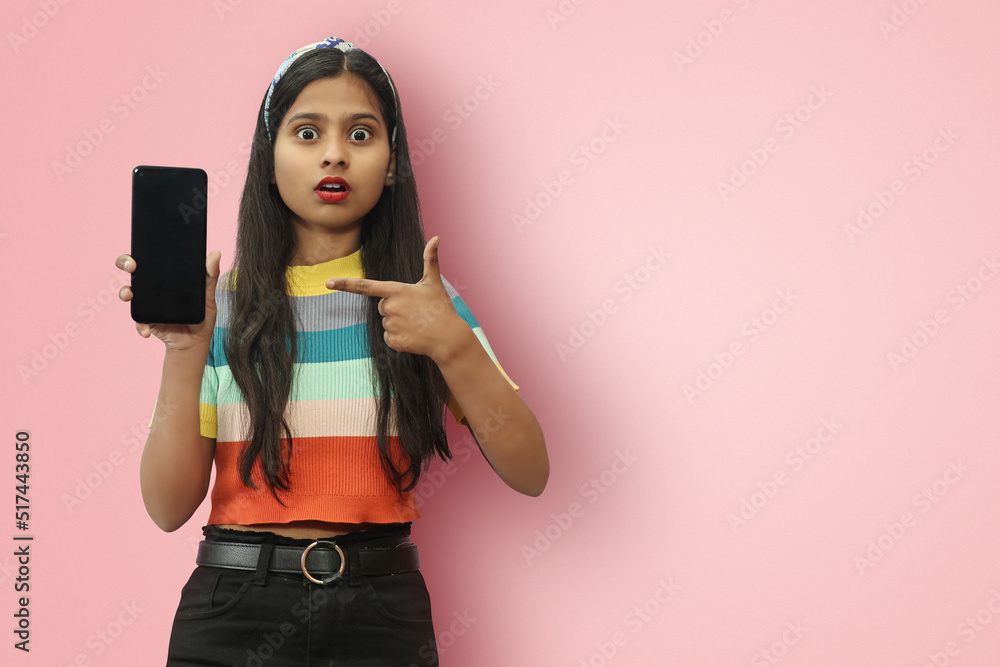 Young Indian Asian girl posing isloated Showing Phone with Blank Screen and Pointing on It to recommend buy after using the product with empty space for text. Young Indian Asian girl posing isloated Showing Phone with Blank Screen and Pointing on It to recommend buy after using the product with empty space for text.