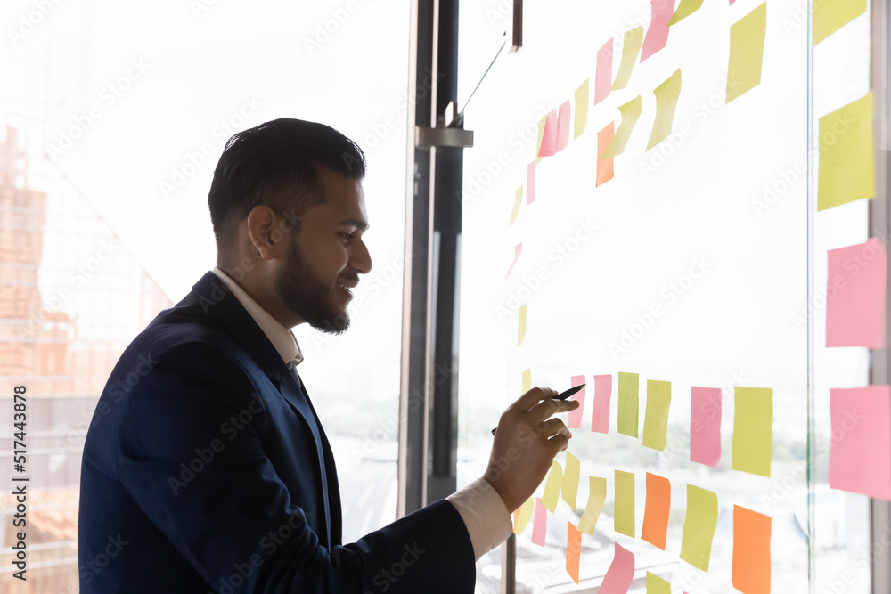 Happy busy Indian businessman making project schedule, writing memos, tasks, ideas on paper sticky notes on glass board, working on startup plan, strategy, smiling. Side view Happy busy Indian businessman making project schedule, writing memos, tasks, ideas on paper sticky notes on glass board, working on startup plan, strategy, smiling. Side view