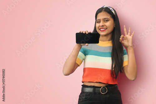Winking young indian asian girl posing isolated showing blank screen smart pone and OK sign. Digital advertisement female model showing the message or new app on cellphone. Mock up copy space.
