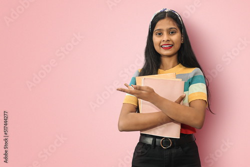 Smiling young indian asian girl student posing islolated holding textbooks and pointing to copyspace with palms up, looking directly at camera, dark haired female expressing positive emotions.
