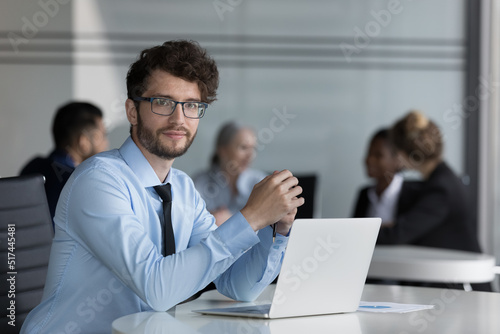 Confident handsome young professional man portrait at laptop in open office space with diverse business group in background. Millennial businessman in glasses sitting at corporate workplace