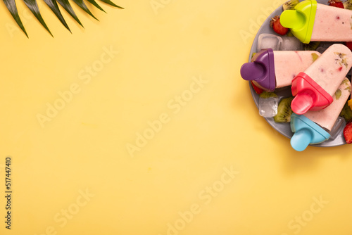 Homemade ice cream from molds on plate with ice and pieces of fruit on colored background and palm leaves with copy space. Top view. Summer food background