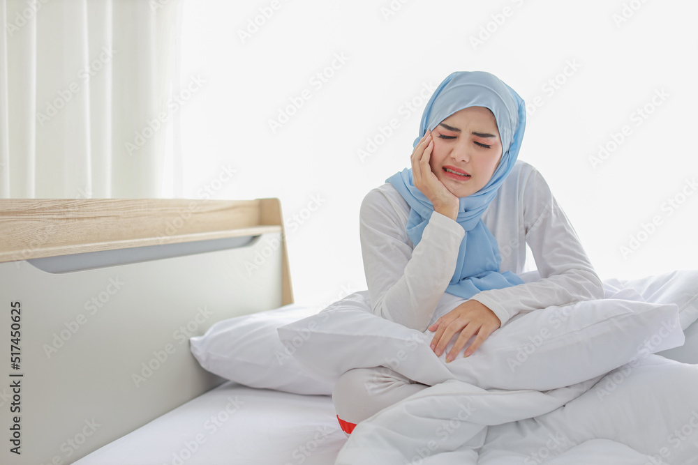 Beautiful and young asian woman in muslim sleepwear with tooth pain emotion sitting on bed. Cute girl touching cheek with her hands and feeling painful from toothache. Dental care and healthy concept. Beautiful and young asian woman in muslim sleepwear with tooth pain emotion sitting on bed. Cute girl touching cheek with her hands and feeling painful from toothache. Dental care and healthy concept.