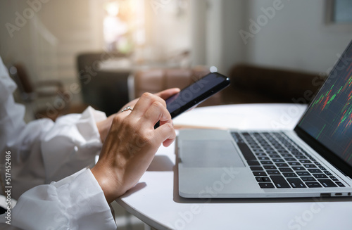 Young asian businesswoman using mobile phone to find information for investment analysis and analyze stock graph on laptop screen to make profitable plans. Close up view