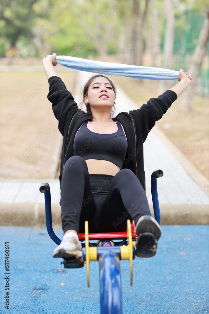 Obraz premium Happy smiling young beautiful asian woman in sportswear relaxing on public exercise machine. Cute and sporty girl working out on bike and using mobile smartphone outdoor