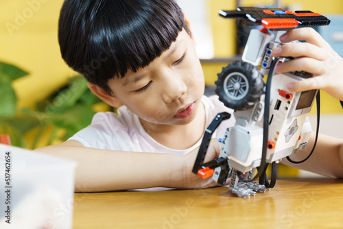 Billede på lærred Smart looking Asian student kid assembling robot, coding and solving engineering problem at home with yellow wall in background