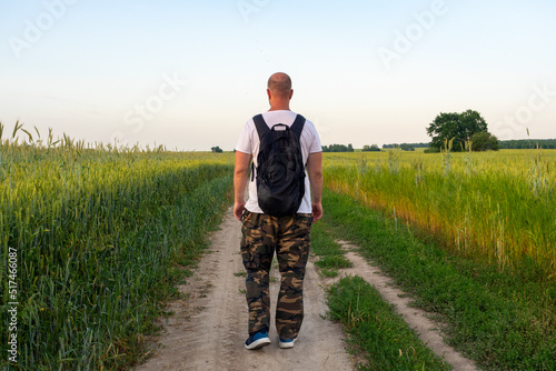 a man walks along a path in a field with a black backpack with his back to the camera. Evening landscape, the concept of travel, recreation, hiking, new discoveries