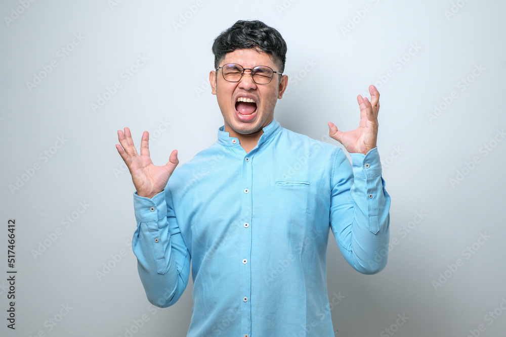 Young asian man wearing casual shirt crazy and mad shouting and yelling with aggressive expression and arms raised. Frustration concept. Young asian man wearing casual shirt crazy and mad shouting and yelling with aggressive expression and arms raised. Frustration concept.