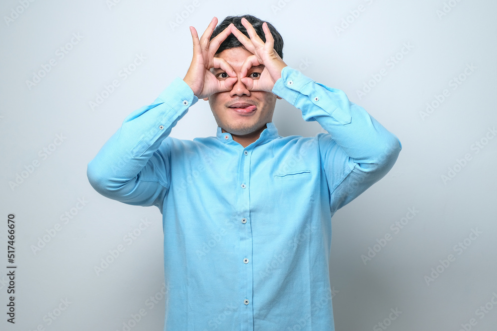 Young handsome man wearing casual shirt doing ok gesture like binoculars sticking tongue out, eyes looking through fingers. Young handsome man wearing casual shirt doing ok gesture like binoculars sticking tongue out, eyes looking through fingers.