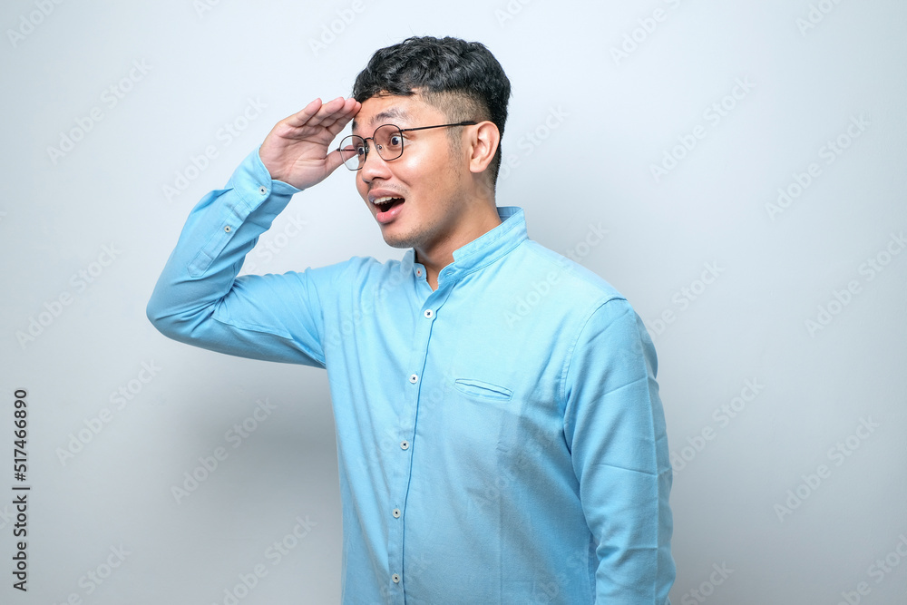 Young asian man wearing casual shirt and glasses looks very happy and smiling looking far away with hand over head Young asian man wearing casual shirt and glasses looks very happy and smiling looking far away with hand over head