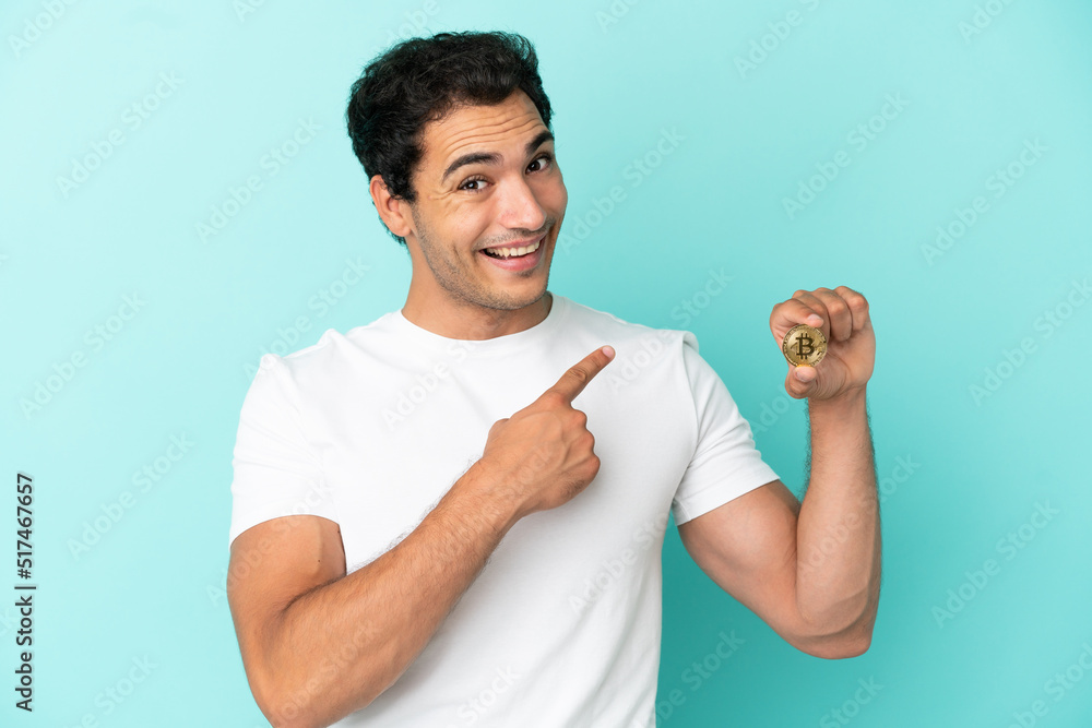 Young man holding a Bitcoin over isolated blue background pointing back
