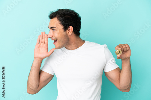 Young man holding a Bitcoin over isolated blue background shouting with mouth wide open to the side