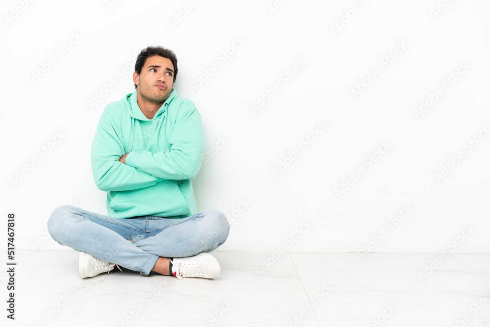 Caucasian handsome man sitting on the floor making doubts gesture while lifting the shoulders Caucasian handsome man sitting on the floor making doubts gesture while lifting the shoulders