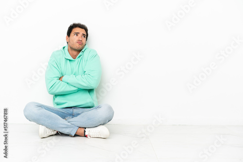 Caucasian handsome man sitting on the floor making doubts gesture while lifting the shoulders