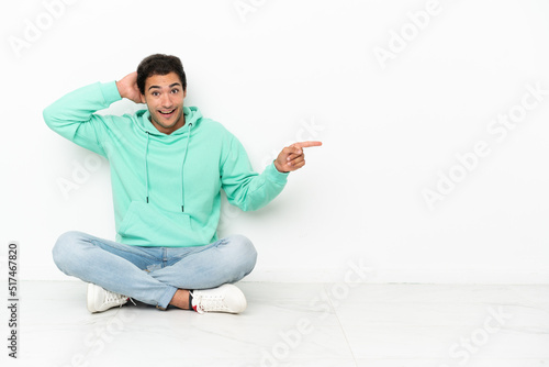 Caucasian handsome man sitting on the floor surprised and pointing finger to the side