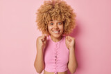Excited curly haired celebrates something triumphs over something bites lips awaits for something wears top poses against pink background winning and rejoicing await for announcement of results