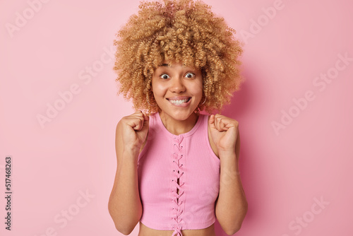 Excited curly haired celebrates something triumphs over something bites lips awaits for something wears top poses against pink background winning and rejoicing await for announcement of results