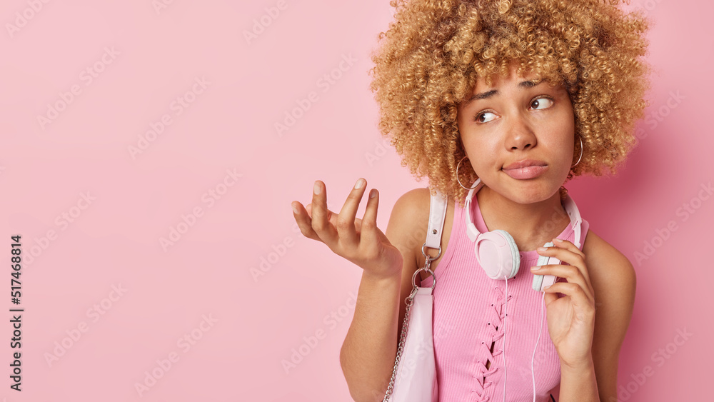 Hesitant clueless young woman with curly hair shrugs shoulders feels confused carries bag wears headphones around neck dressed in t shirt isolated over pink background copy space for your promo