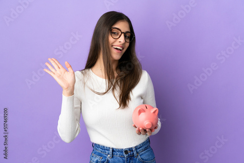 Young caucasian woman holding a piggybank isolated on purple background saluting with hand with happy expression