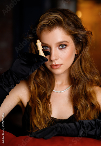A beautiful Caucasian girl in a black dress poses with a chess piece. Fashion shooting