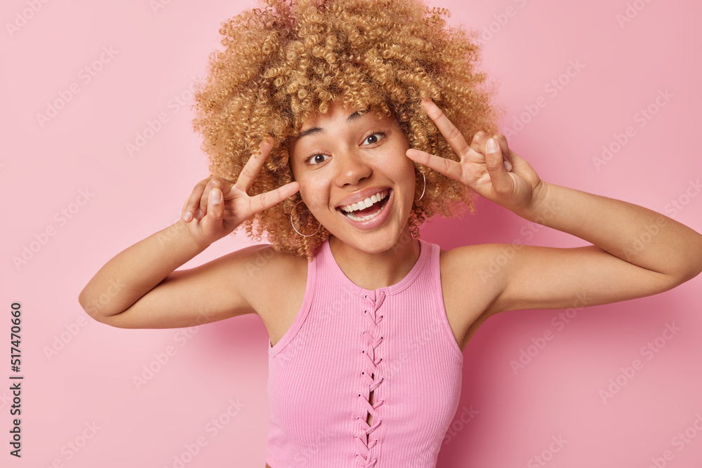 Positive curly haired woman makes peace gesture smiles happily at camera looks carefree enjoys life dressed in t shirt poses against pink background expresses joy and optimism. Salute v sign Positive curly haired woman makes peace gesture smiles happily at camera looks carefree enjoys life dressed in t shirt poses against pink background expresses joy and optimism. Salute v sign