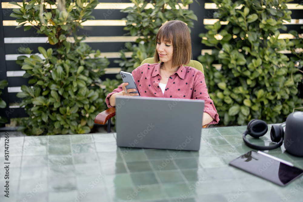 Young woman works on laptop and phone at outdoor workspace in the garden. Concept of remote work from comfortable home office or work during vacations Young woman works on laptop and phone at outdoor workspace in the garden. Concept of remote work from comfortable home office or work during vacations