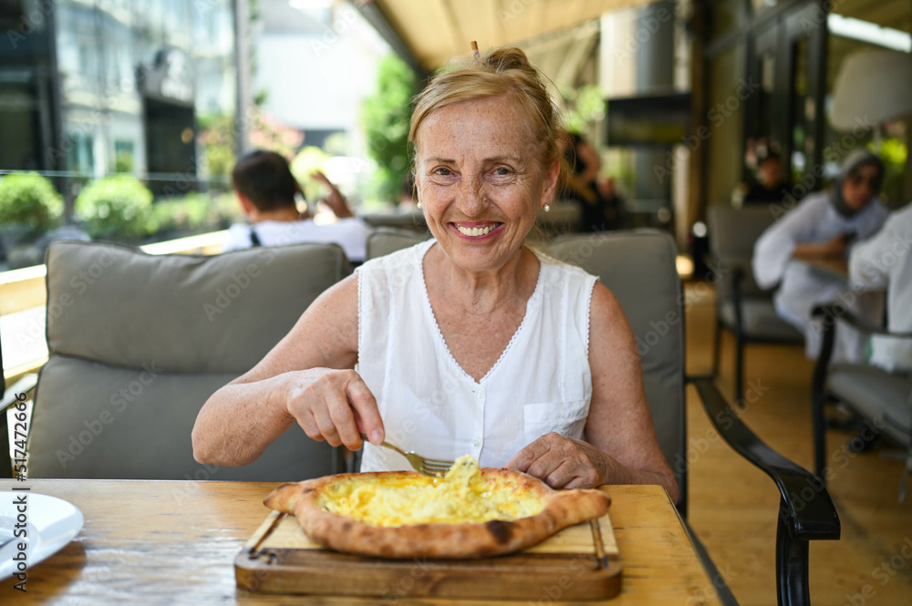 Happy senior woman with perfect smile eating adjarian khachapuri outdoors at summer sidewalk street cafe or restaurant terrace. Retired mature people holiday vacation, active lifestyle concept
