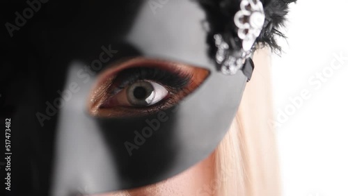 Beauty and fashion concept. Beautiful blonde woman with evening make-up looking through black carnival mask when slowly walks away. Eye is in camera focus