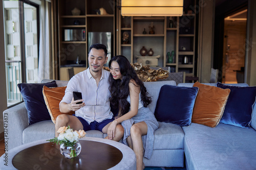 Husband and wife taking selfie photo sitting on sofa. Happy asian couple having fun in living room at home