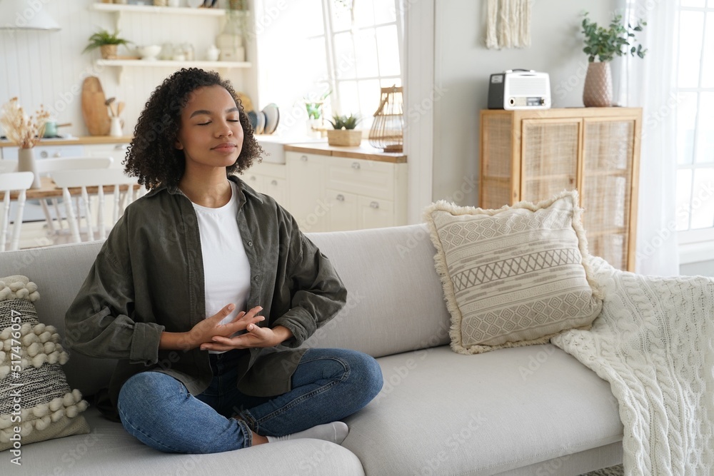Biracial young girl enjoy meditation, practice yoga, sitting on sofa in lotus pose at home Biracial young girl enjoy meditation, practice yoga, sitting on sofa in lotus pose at home