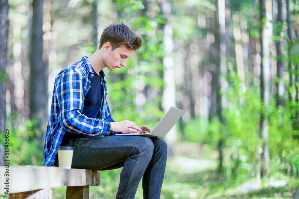 A young man is typing on a laptop in a summer forest. A young man is typing on a laptop in a summer forest.
