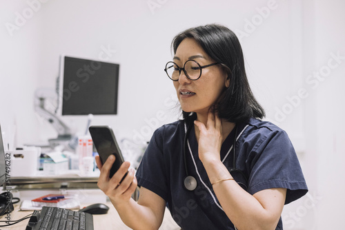 Female otolaryngologist explaining while touching throat during online consultation through smart phone in medical clini photo