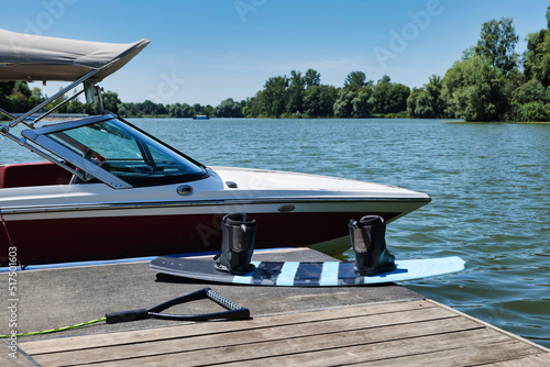 Wakeboard with rope and handle lying next to a ski boat, wakeboard boat which is docked at the pier. A calm and empty lake in the background. Adventure, leisure, action sports scene. 