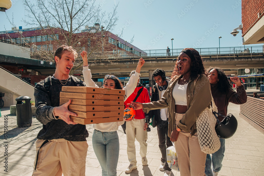Man carrying stack of pizza boxes while walking with friends at street on sunny day Man carrying stack of pizza boxes while walking with friends at street on sunny day