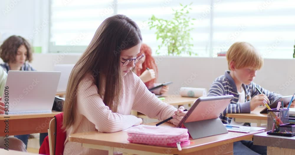 Caucasian schoolgirl in glasses sitting at desk in classroom, writing and using tablet device for study. Time at school. Smart girl learning at lesson and checking exercise in copybook with computer. Caucasian schoolgirl in glasses sitting at desk in classroom, writing and using tablet device for study. Time at school. Smart girl learning at lesson and checking exercise in copybook with computer.