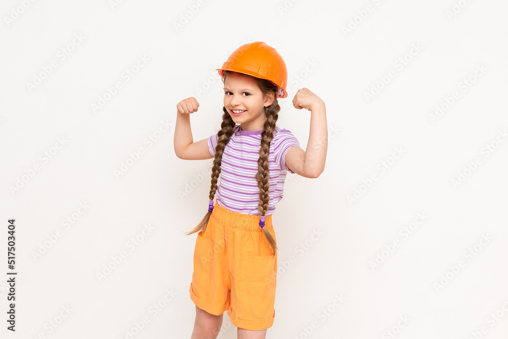 A little girl in a construction helmet and pigtails shows the strength of her biceps on a white isolated background. The profession of an engineer for a child. A little girl in a construction helmet and pigtails shows the strength of her biceps on a white isolated background. The profession of an engineer for a child.